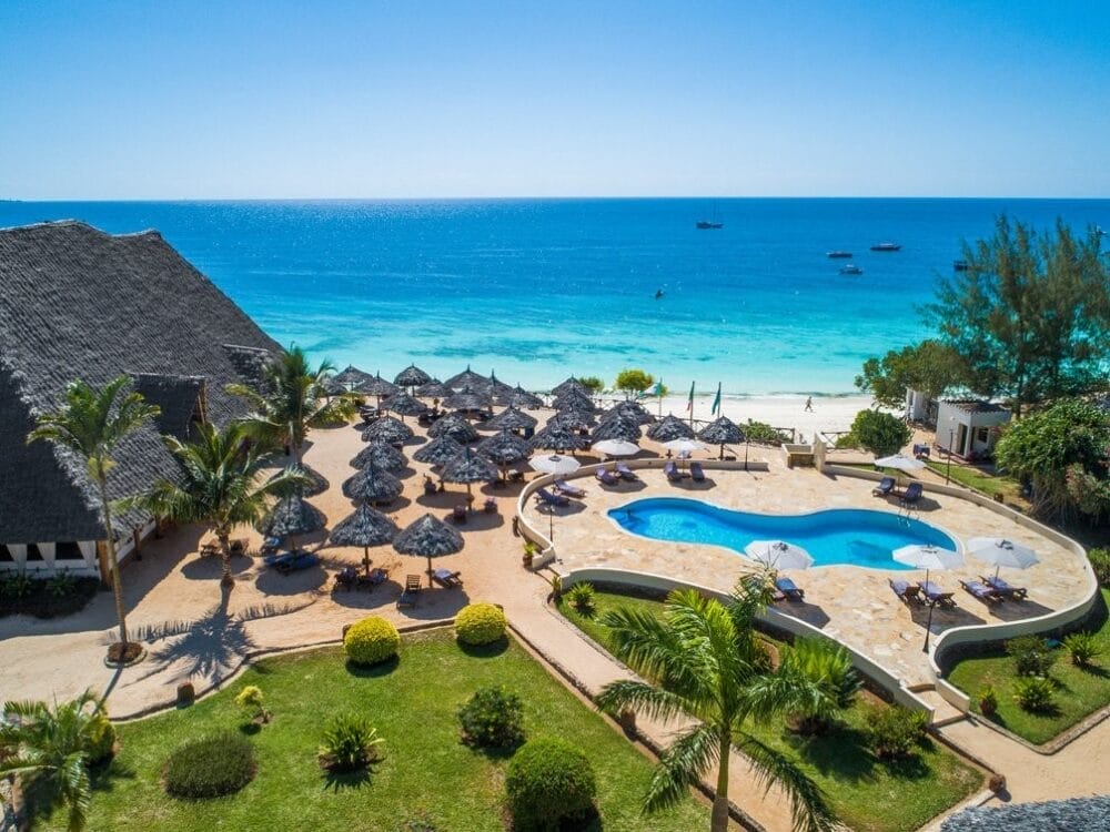 Vista aerea del Veraclub Sunset Beach, un resort tropicale con una grande piscina, palme, ombrelloni di paglia, sedie a sdraio e giardini che si affacciano sull'oceano turchese sotto un cielo azzurro e limpido.