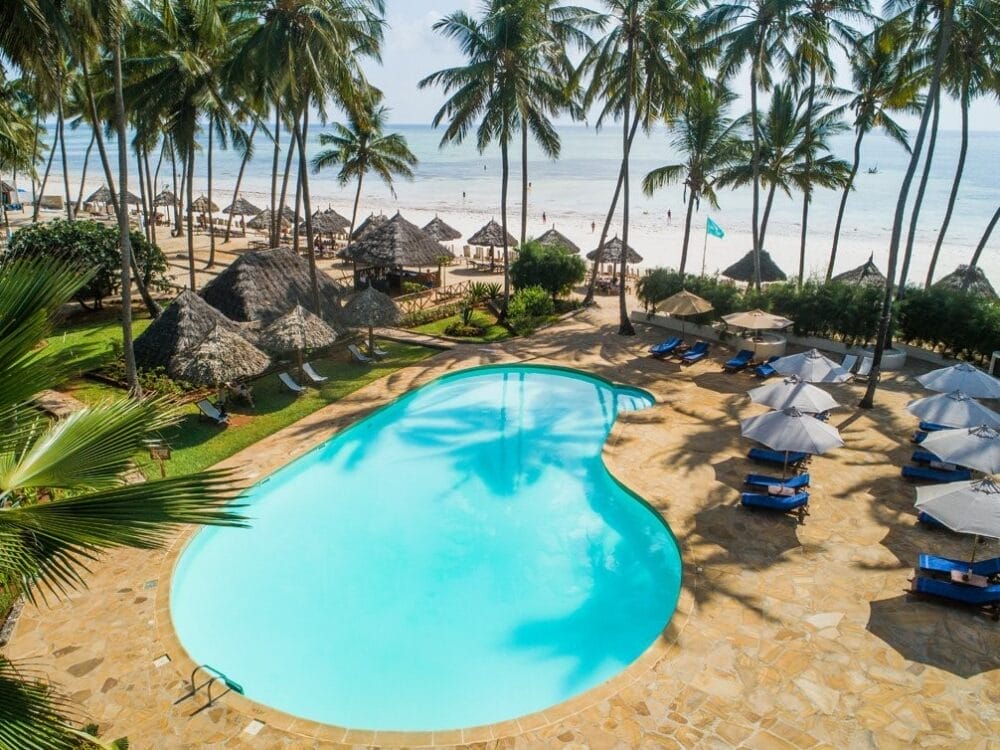 Un resort tropicale come il Veraclub Zanzibar presenta una piscina a forma di rene circondata da sedie a sdraio, ombrelloni, palme, capanne di paglia e una spiaggia sabbiosa con persone e onde sullo sfondo.