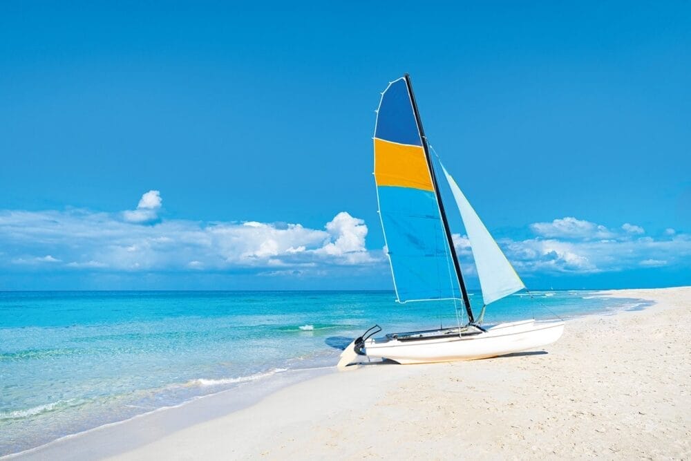 Una piccola barca a vela con vele blu e arancioni riposa su una spiaggia sabbiosa in un'acqua limpida e turchese a Varadero, vicino al resort Veraclub Starfish, sotto un cielo blu brillante con nuvole sparse.