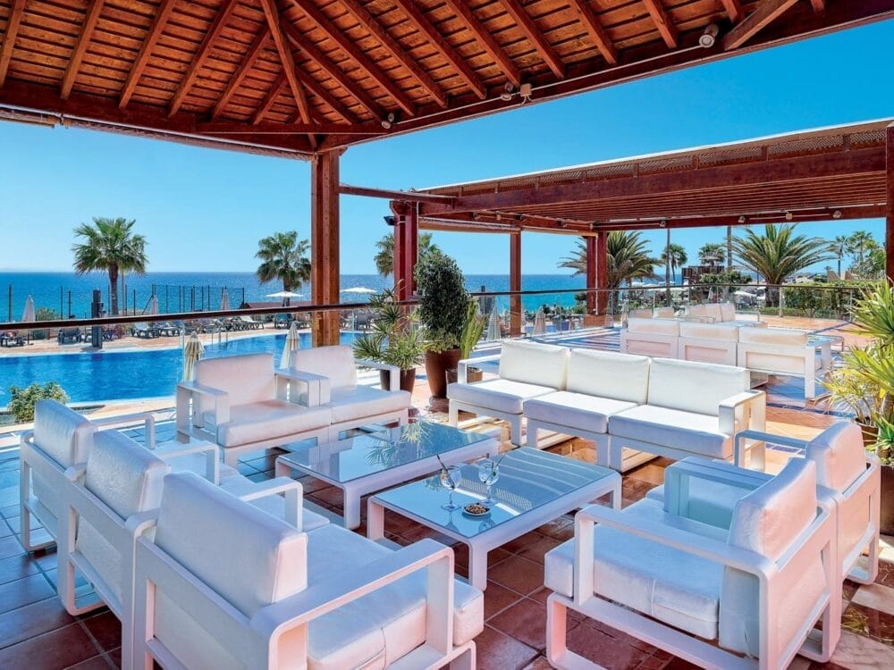 La lounge coperta all'aperto del Veraclub Tindaya è caratterizzata da divani bianchi e tavoli in vetro che si affacciano sulla piscina, sulle palme e sull'oceano sotto un cielo azzurro e limpido, offrendo un'atmosfera rilassata da resort tropicale all inclusive a Fuerteventura.