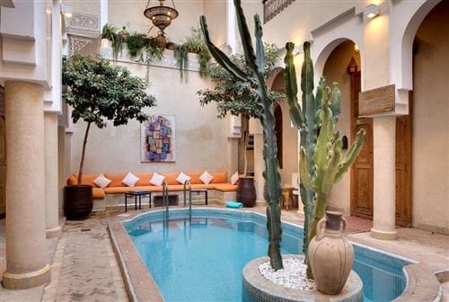 L'accogliente cortile dell'Angsana Riads di Marrakech è caratterizzato da una piccola piscina, cactus e alberi in vaso. Una lunga panca arancione con cuscini, porte ad arco e decorazioni marocchine creano un alloggio di lusso immerso nella calda luce naturale.