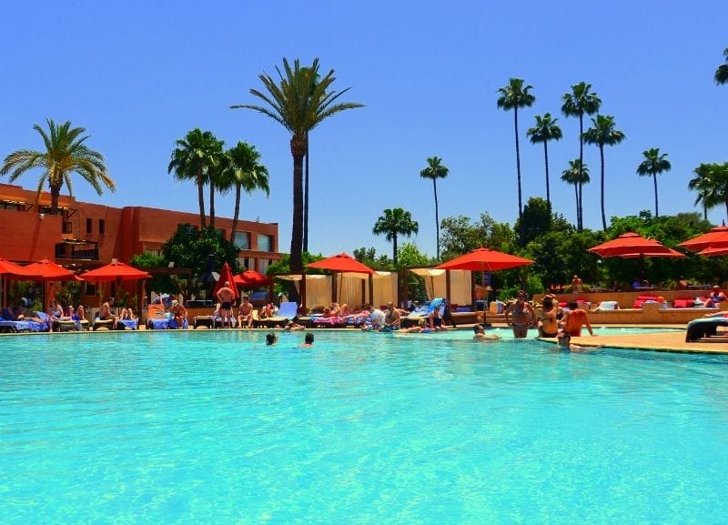 Persone si rilassano presso la grande piscina all'aperto dei Giardini Medina, circondata da palme, ombrelloni rossi e lettini in una giornata di sole. Sullo sfondo, l'edificio Kappa City color terracotta.