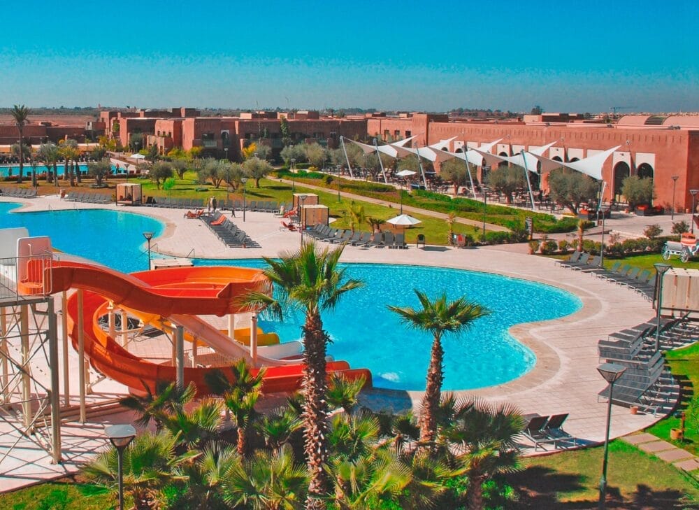Grande resort all'aperto al Kenzi Agdal Medina con piscine curve, scivoli d'acqua, palme, sedie a sdraio e posti a sedere all'ombra sotto baldacchini di vele bianche, con gli edifici marroni dell'hotel Kenzi e un cielo azzurro sullo sfondo.