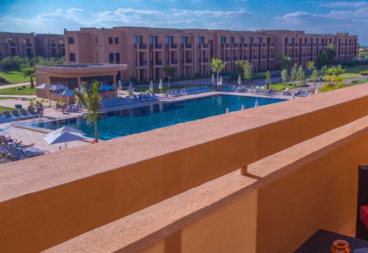 Vista da un balcone che si affaccia sull'area della piscina dell'hotel Aqua Mirage a Marrakech, circondata da lettini, ombrelloni, palme e un moderno edificio a più piani sotto un cielo azzurro e soleggiato.