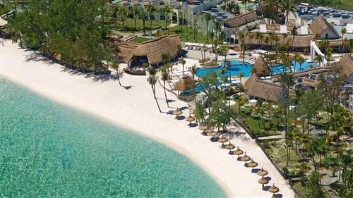 Vista aerea dell'Ambre Sun Resort, un rifugio tropicale sulla spiaggia con sabbia bianca, acqua turchese, lettini e ombrelloni lungo la riva, palme, piscine ed edifici con tetti di paglia circondati da una vegetazione lussureggiante.