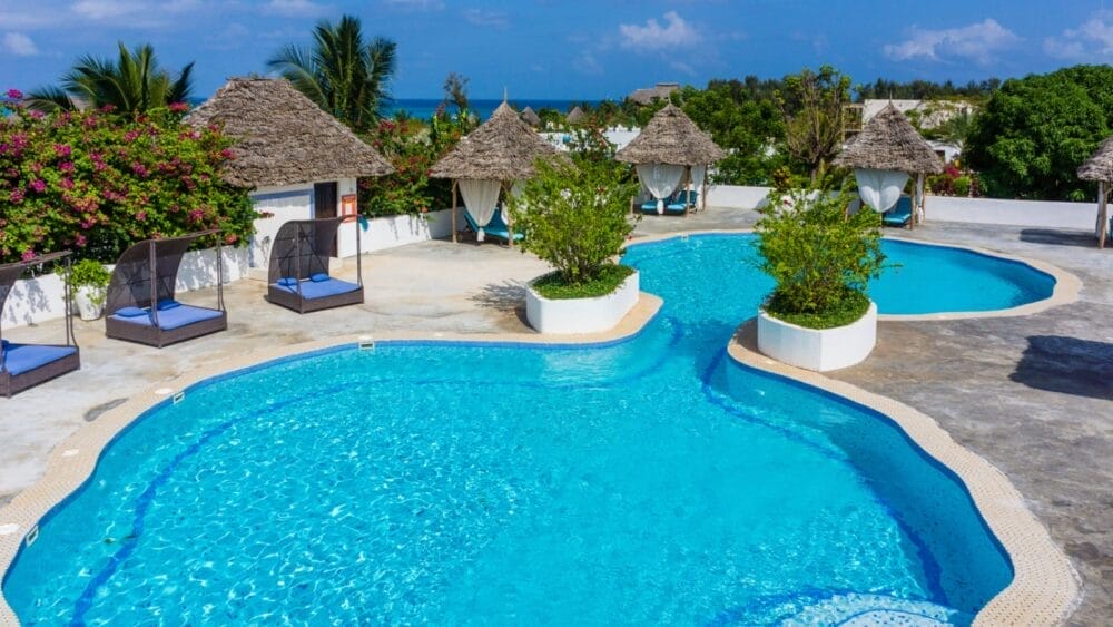 Piscina all'aperto con acqua cristallina al Bravo Kendwa Beach Resort, circondata da piante tropicali, sedie a sdraio e cabine con tetto di paglia. L'area soleggiata della piscina offre una vista sugli alberi e sull'oceano scintillante sullo sfondo.