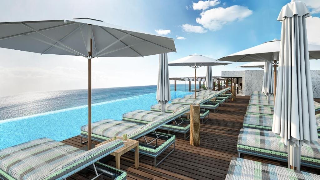 Piscina a sfioro sul tetto con sedie a sdraio e ombrelloni su un ponte di legno dell'Ocean V Hotel, con vista sull'oceano sotto un cielo blu brillante con nuvole sparse.