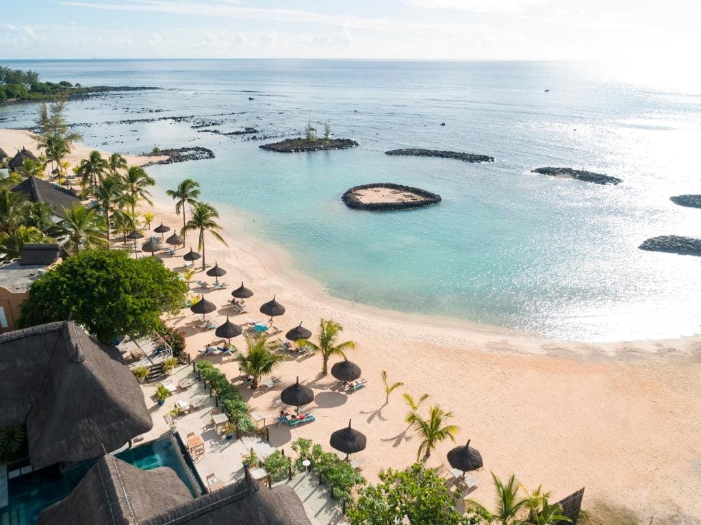 Vista aerea del Veranda Pointe Aux Biches, uno splendido resort sulla spiaggia di Mauritius con acque cristalline, arenile sabbioso, palme, ombrelloni con tetto di paglia e piccole isole rocciose che punteggiano il mare calmo vicino agli edifici del resort a bordo piscina.