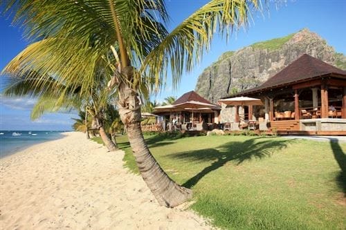 Il Lux Le Morne, un resort tropicale sulla spiaggia, è caratterizzato da palme, ville in legno e posti a sedere all'aperto che si affacciano sulla sabbia bianca e sull'acqua cristallina, con una maestosa montagna rocciosa che si erge sullo sfondo sotto un cielo blu brillante.