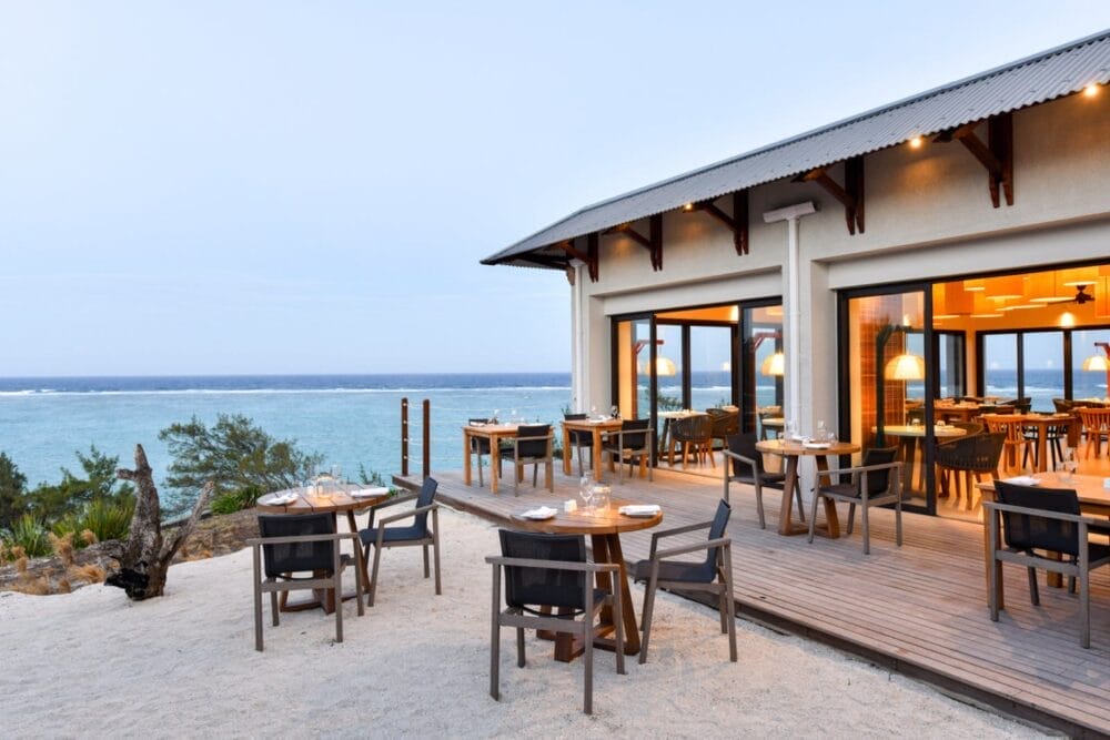 Ristorante all'aperto del Constance Tekoma Rodrigues con tavoli e sedie in legno sulla sabbia e una terrazza, con vista sull'oceano. Le grandi finestre rivelano l'interno illuminato da una luce calda. Gli alberi e il cielo blu creano uno sfondo tranquillo.