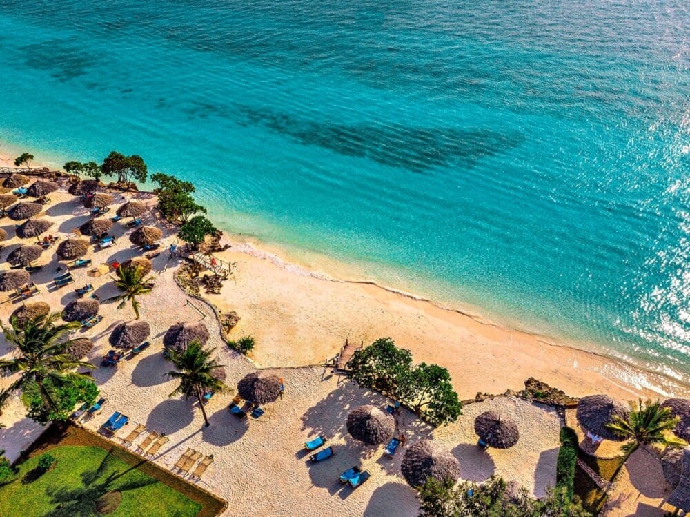 Vista aerea del Bravo Premium Sandies Baobab, una spiaggia tropicale con acqua turchese e trasparente, sabbia dorata, lettini e ombrelloni di paglia. Palme e vegetazione lussureggiante fanno ombra lungo il litorale.