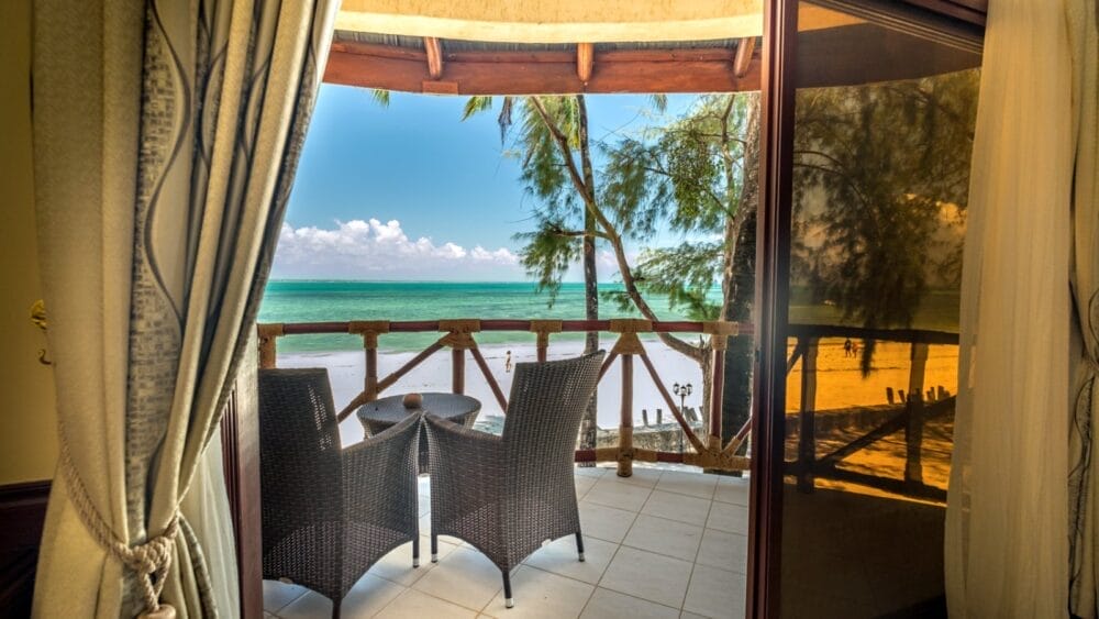 Vista da una camera del Club Coralia Zanzibar Resort attraverso tende aperte e una porta a vetri su un balcone con sedie in vimini, che si affaccia sulla spiaggia sabbiosa di Kae Beach, sul mare turchese, sugli alberi ondeggianti e su un cielo blu brillante.