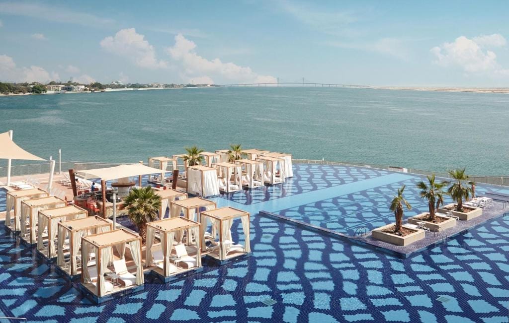 L'area esterna della piscina dell'hotel di lusso è caratterizzata da cabine, palme e piastrelle con motivi blu che si affacciano su un mare calmo sotto un cielo limpido. La terra e un ponte sono visibili in lontananza al Royal M Hotel Abu Dhabi.