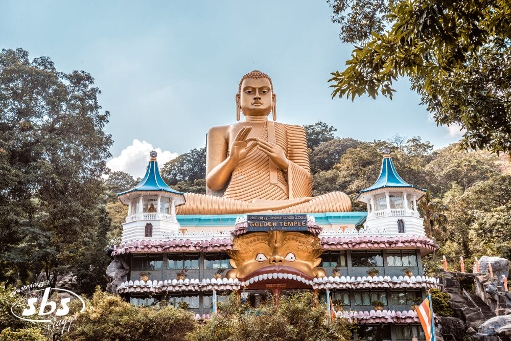 Una grande statua dorata di Buddha si trova in cima al colorato Tempio d'Oro di Dambulla, nello Sri Lanka, circondato da alberi rigogliosi e dal cielo azzurro. L'ingresso del tempio presenta una bocca di leone decorata e un'architettura automatica Bozza.
