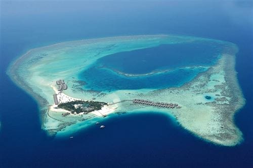 Veduta aerea di Constance Moofushi alle Maldive, una piccola isola tropicale circondata da acque turchesi e barriere coralline, caratterizzata da gruppi di edifici e bungalow overwater con il blu intenso dell'oceano.