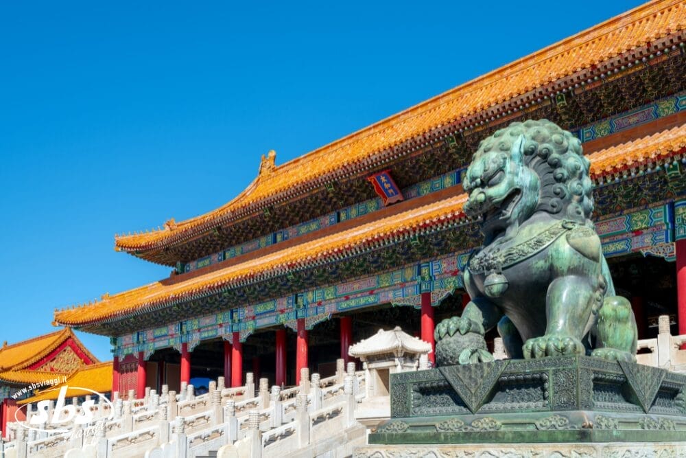 La statua di un leone di bronzo si trova davanti a un edificio colorato e decorato, con un'architettura tradizionale cinese e un tetto arancione brillante, sotto un cielo azzurro e limpido: una scena suggestiva che ricorda un'artistica Bozza automatica.