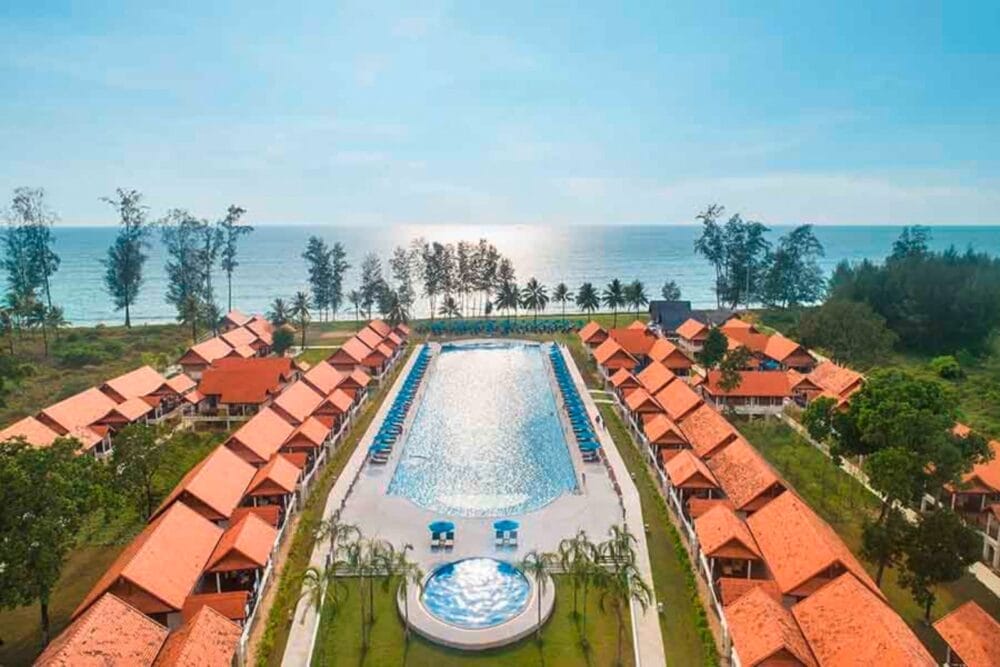 Una grande piscina all'aperto è circondata da file di ville Bravo Premium di Khao Lak dai tetti rossi, con palme, prati verdi e una vista sull'oceano sullo sfondo sotto un cielo azzurro e limpido.