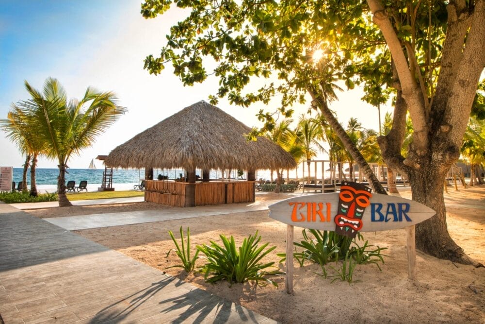 Una spiaggia tropicale con palme, un tiki bar con tetto in paglia e un'insegna in legno con la scritta "Tiki Bar" e una maschera tiki colorata. La luce del sole filtra attraverso gli alberi del Bravo Viva Dominicus Beach, mentre l'oceano è visibile sullo sfondo.