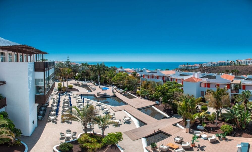 Il Bravo Fuerteventura è un resort con edifici bianchi, tetti di tegole arancioni, palme, piscine multiple e sedie a sdraio, che si affaccia sull'oceano sotto un cielo azzurro e limpido.