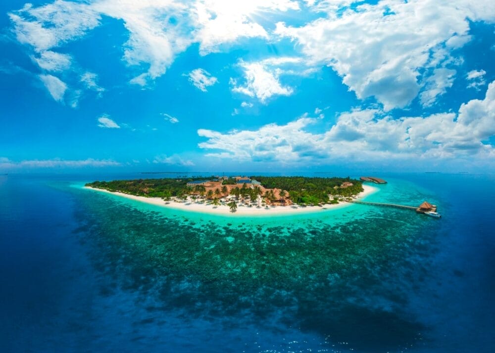 Un'isola tropicale circondata da un oceano limpido e dalla barriera corallina, con una vegetazione lussureggiante e spiagge sabbiose sotto un cielo luminoso e parzialmente nuvoloso. Un molo si estende nell'acqua, catturando il fascino sereno di un resort delle Maldive.