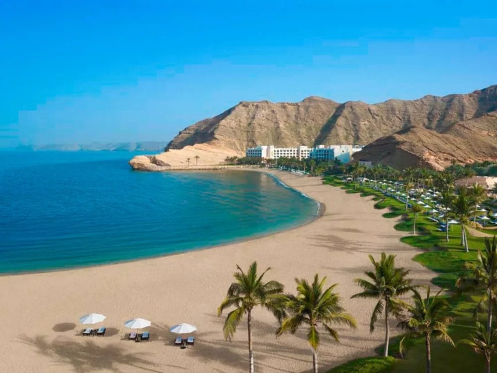 Una spiaggia sabbiosa con palme, ombrelloni bianchi e sedie a sdraio costeggia il mare blu dello Shangri-La Barr Al Jissah. Sullo sfondo, il resort Al Waha è incastonato alla base di aspre montagne sotto un cielo azzurro e limpido.