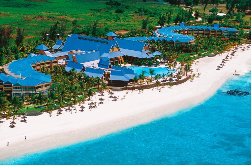 Vista aerea del Victoria Beachcomber Resort & Spa, che mostra edifici dai tetti blu, una grande piscina, palme, lettini e ombrelloni lungo una spiaggia di sabbia bianca accanto all'acqua turchese dell'oceano.