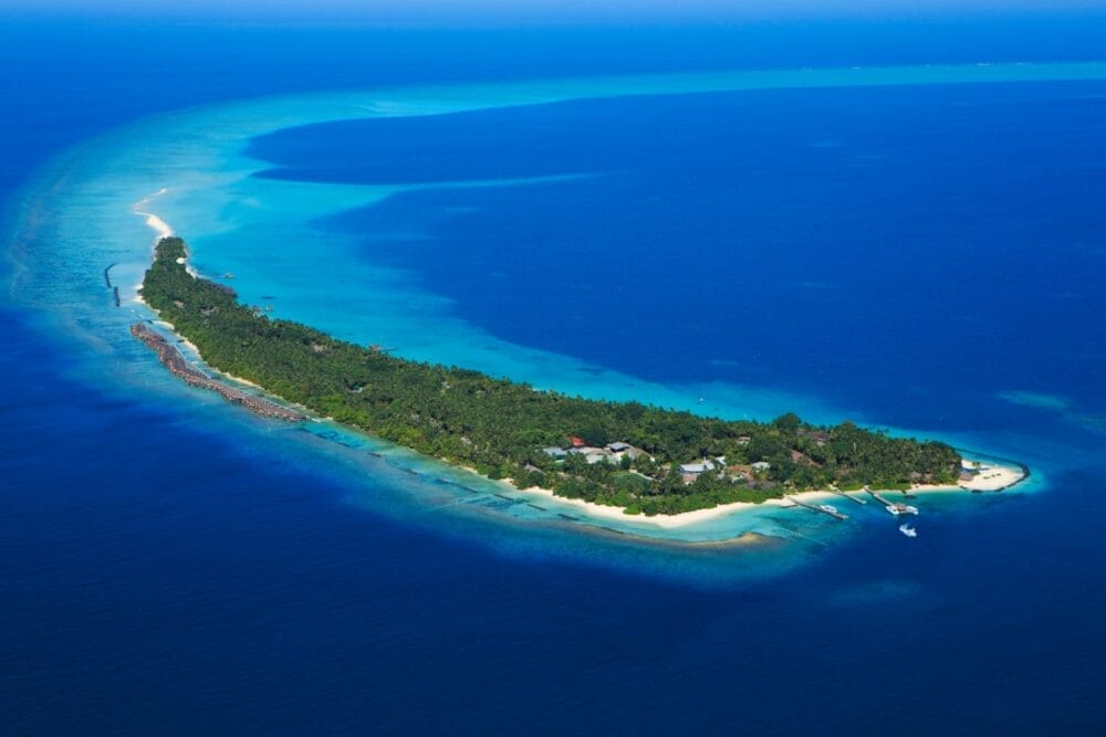 L'isola di Kuramathi, lussureggiante e verde, circondata da acque turchesi e spiagge di sabbia bianca, vista dall'alto. Diverse barche sono ancorate vicino alla riva e gli edifici dei resort delle Maldive fanno capolino tra gli alberi.