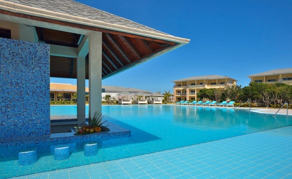 Una grande piscina all'aperto con bar e sgabelli al Paradisus Varadero, circondata da sedie a sdraio e da moderni edifici del resort sotto un cielo azzurro e limpido. Una composizione di frutta è collocata a bordo piscina.