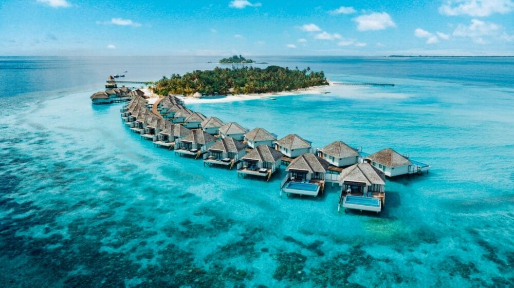 Vista aerea dei bungalow overwater disposti a curva sopra le acque turchesi di un resort delle Maldive, che conducono a una piccola isola lussureggiante con palme, circondata dall'oceano blu sotto un cielo soleggiato.