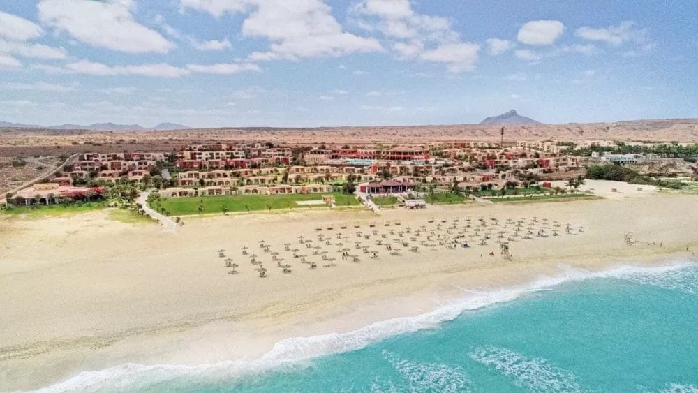Vista aerea del Veraclub a Boa Vista Beach, con file di ombrelloni e sedie a sdraio sulla sabbia dorata, onde oceaniche turchesi e un paesaggio desertico con montagne sullo sfondo sotto un cielo parzialmente nuvoloso.