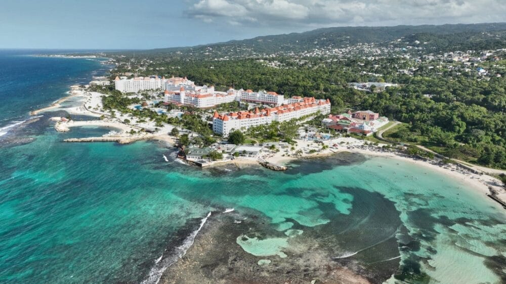 Vista aerea del Bahia Principe Grand Jamaica, un hotel all-inclusive con edifici dai tetti rossi immersi in una vegetazione lussureggiante, spiagge di sabbia bianca e acque turchesi lungo una costa tropicale. Colline e una città vicina completano la scena.
