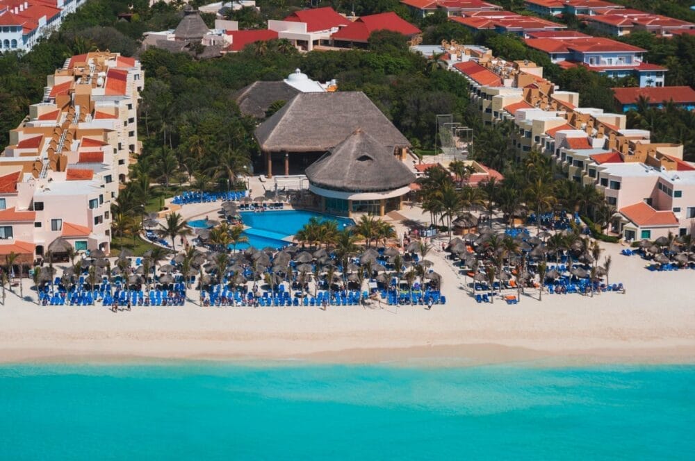 Vista aerea del resort Wyndham Viva Maya con piscina, edificio con tetto in paglia, file di sedie a sdraio e ombrelloni blu sulla spiaggia sabbiosa e acqua turchese dell'oceano, circondato da palme ed edifici colorati.