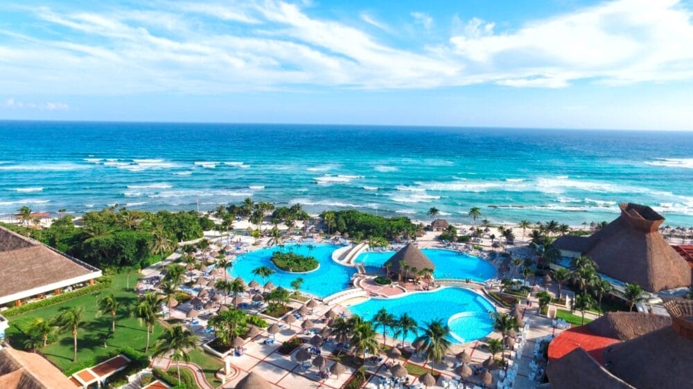 Vista aerea del resort Grand Tulum con grandi piscine blu, palme, lettini, edifici con tetti di paglia e onde turchesi dell'oceano sotto un cielo parzialmente nuvoloso.