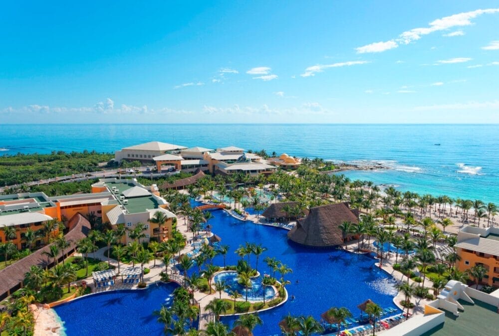 Vista aerea del Barcelo Maya Grand Resort con grandi piscine blu, palme ed edifici vicino alla costa, sullo sfondo dell'oceano e del cielo azzurro.