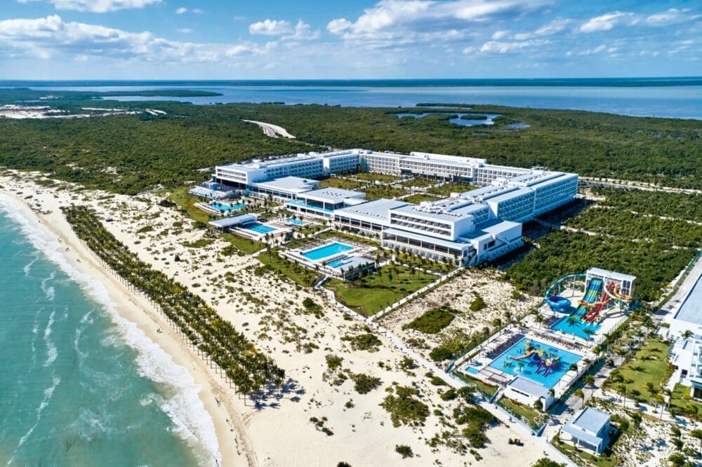 Vista aerea del Riu Palace Costa Mujeres, uno splendido hotel all-inclusive con diverse piscine, un parco acquatico e palme lungo una spiaggia sabbiosa accanto all'oceano turchese e alla vegetazione lussureggiante sotto un cielo parzialmente nuvoloso.