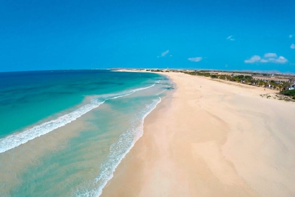 Veduta aerea di Bravo Praia De Chaves, una lunga spiaggia sabbiosa con le onde turchesi dell'oceano che si infrangono dolcemente a riva sotto un cielo azzurro e limpido. La costa si estende in lontananza con una vegetazione rada e piccoli edifici visibili sulla destra.