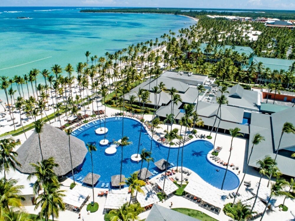 Vista aerea del Barceló Bavaro Beach, un resort all inclusive a Punta Cana, caratterizzato da una grande piscina sinuosa, lettini, palme ed edifici vicino all'acqua turchese dell'oceano e alla spiaggia sabbiosa sotto un cielo azzurro e limpido.