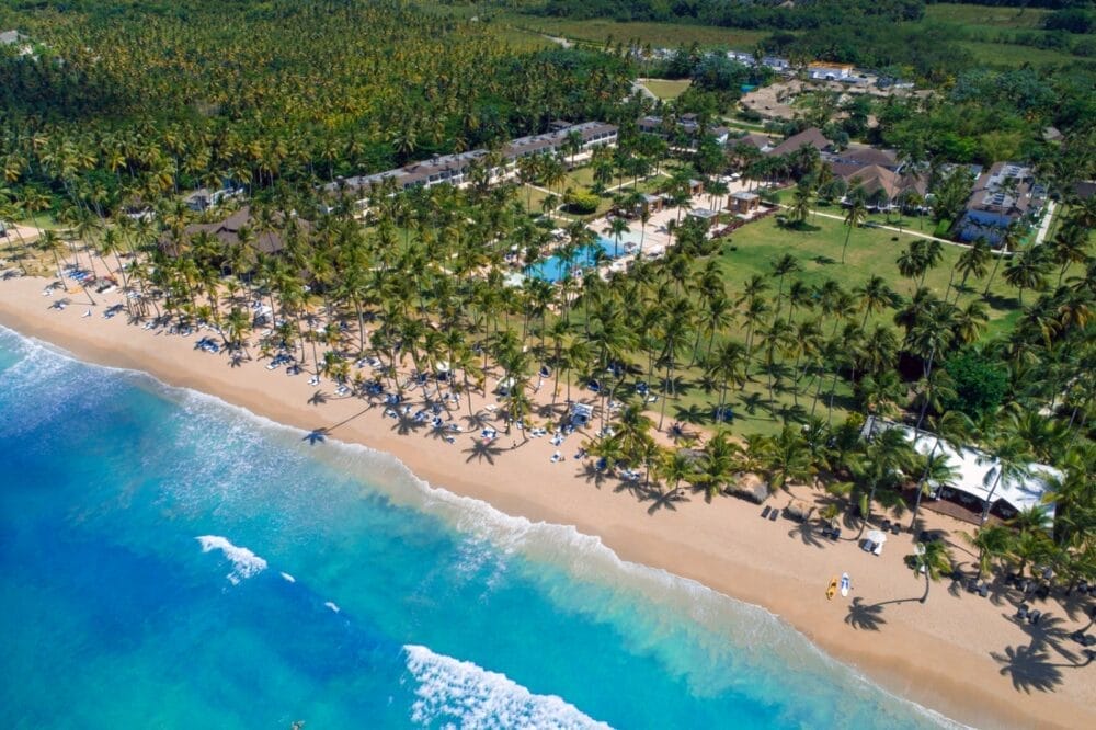 Vista aerea della spiaggia tropicale del Viva V Samana, con acque turchesi e trasparenti, onde dolci e sabbia dorata fiancheggiata da palme. Gli edifici, le piscine e il verde lussureggiante di Wyndham sono visibili più all'interno mentre le persone si rilassano lungo la riva.