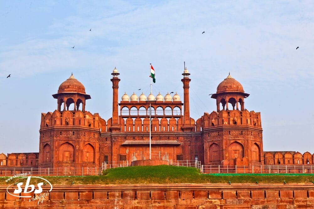 Il Forte Rosso di Delhi, India Splendida, con la bandiera indiana che sventola in cima alla sua cupola centrale, si staglia contro un cielo blu con gli uccelli che svettano sopra e l'erba verde in primo piano.