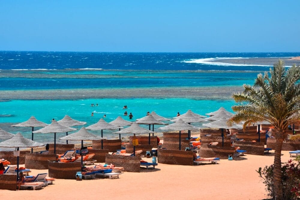 File di ombrelloni di paglia e sedie a sdraio costeggiano la spiaggia sabbiosa del Bravo Fantazia Resort, affacciata su un mare blu turchese. La gente nuota in mare, mentre una palma ondeggia sulla destra sotto un cielo limpido e soleggiato.