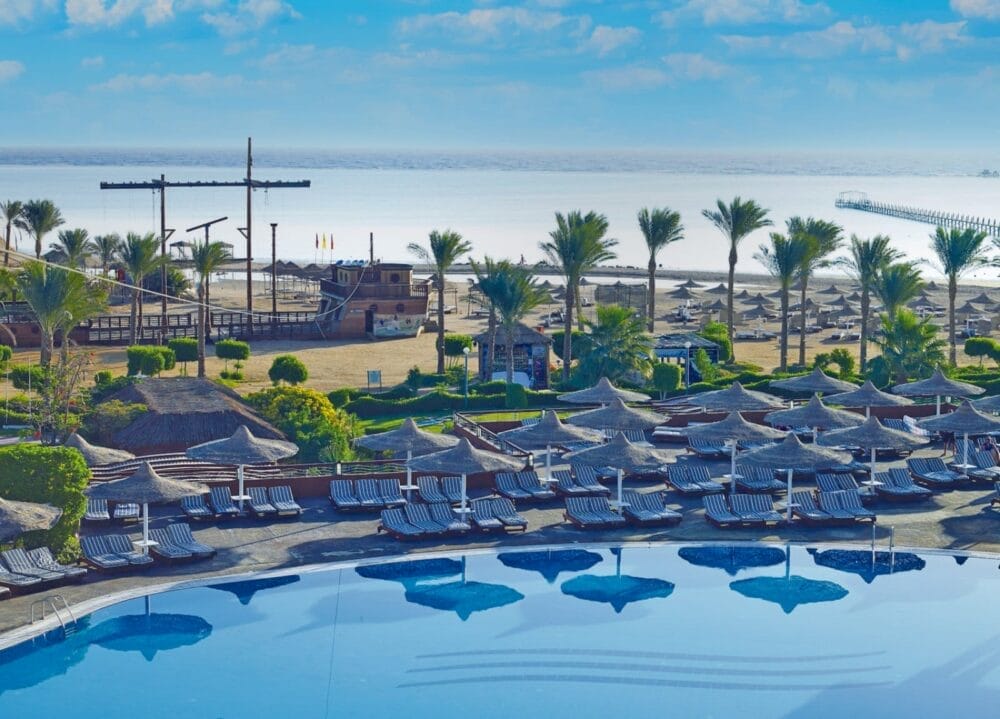 La piscina del Coral Sea Holiday Resort è caratterizzata da file di sedie a sdraio e ombrelloni vuoti, che si affacciano su una spiaggia sabbiosa con palme, strutture ombreggianti di paglia e una grande nave di legno replicata dal mare sotto un cielo blu brillante.