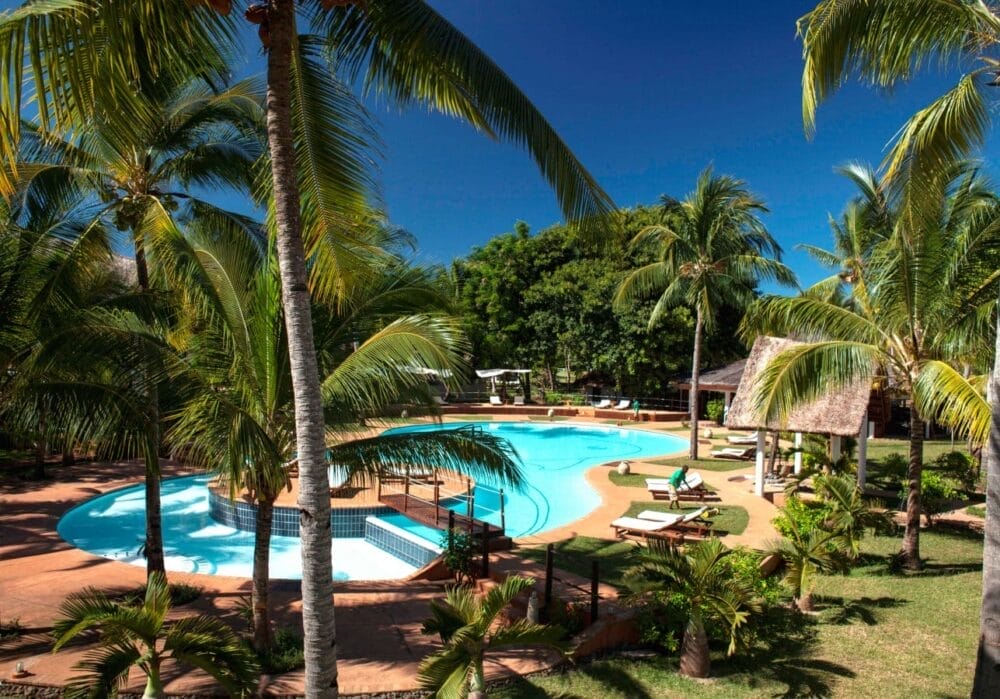 La piscina tropicale dell'Amarina Alpiclub, circondata da palme, lettini e un padiglione con tetto di paglia sotto un cielo azzurro e limpido, crea un'atmosfera di vacanza rilassante e lussureggiante.
