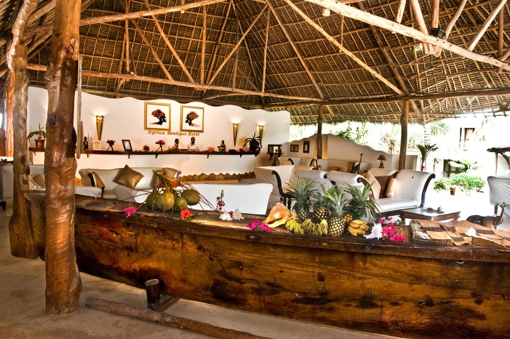 Salone all'aperto del My Blue Hotel con divani bianchi, tavolini in legno e tetto di paglia. Una barca di legno piena di frutta e fiori tropicali funge da centrotavola, circondata da decorazioni rustiche e opere d'arte a parete. La luce naturale riempie lo spazio.