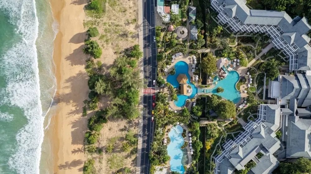 Veduta aerea di una spiaggia con onde, una strada e un resort con diverse piscine circondato da verde ed edifici. La spiaggia, la strada e il resort Bozza automatica corrono paralleli da sinistra a destra.