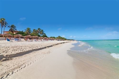 Una spiaggia sabbiosa con acqua turchese, palme e persone che si rilassano sotto gli ombrelloni di paglia a Cuatro Palmas in una giornata di sole con un cielo azzurro e limpido: l'ideale per gli ospiti Over 16 Anni in cerca di serenità.