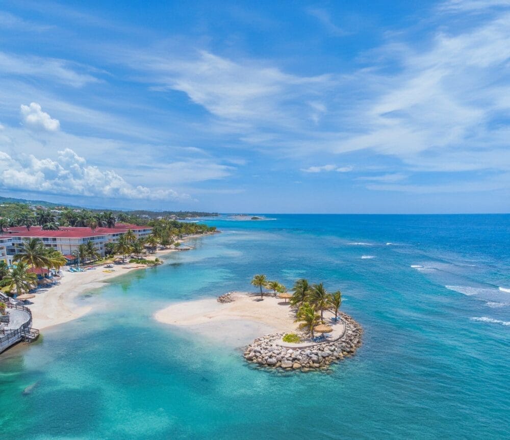 Veduta aerea di una spiaggia tropicale vicino ai resort di Montego Bay, con acqua turchese, palme, spiaggia sabbiosa e un piccolo isolotto roccioso. Gli edifici dei resort e la vegetazione lussureggiante costeggiano la costa sotto un cielo blu con nuvole sparse.
