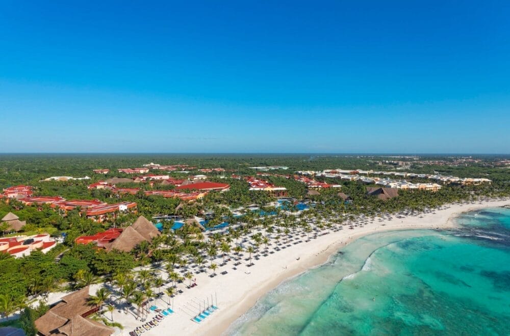 Vista aerea del Barcelo Maya Caribe, un resort sulla spiaggia tropicale con sabbia bianca, acqua turchese, file di sedie a sdraio, palme ed edifici dai tetti rossi circondati da una vegetazione lussureggiante sotto un cielo azzurro e limpido. Prenotate con Alpiselect per soggiorni curati.