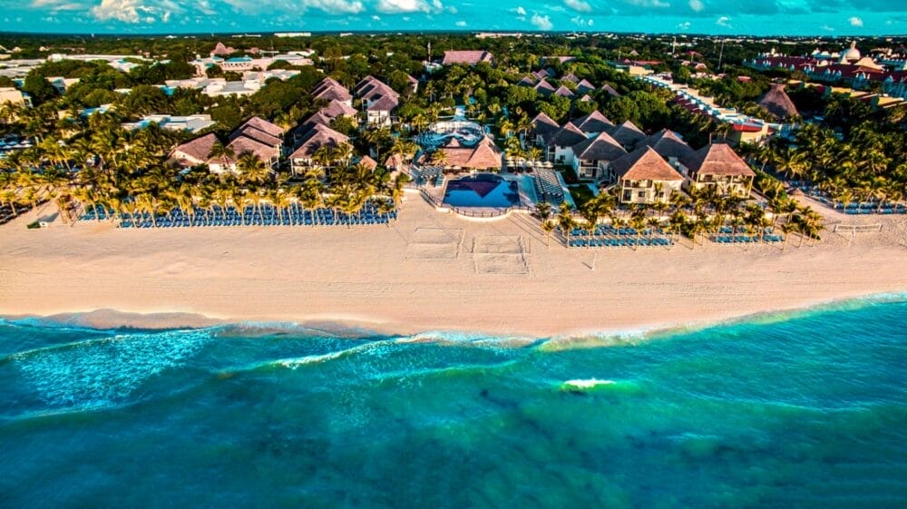 Veduta aerea di Allegro Playacar, un resort all-inclusive a Playa del Carmen, con edifici dai tetti di paglia, palme, sedie a sdraio su sabbia bianca, onde turchesi dell'oceano, vegetazione lussureggiante e una città sullo sfondo sotto un cielo azzurro.