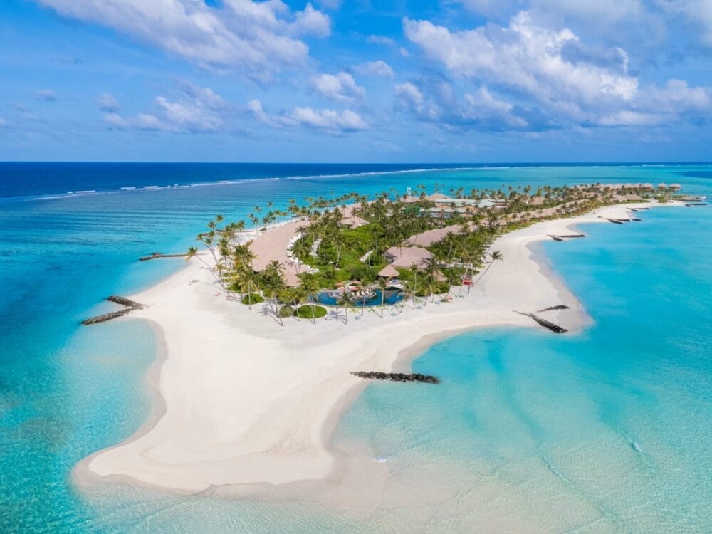 Vista aerea di Barceló Whale Lagoon, un'isola tropicale con spiagge di sabbia bianca, acqua turchese, palme e pochi edifici. L'isola è circondata da un oceano limpido sotto un cielo parzialmente nuvoloso.