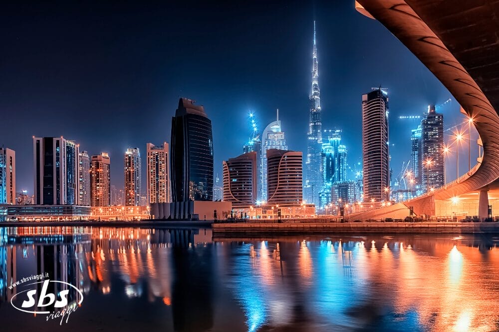 Un vivace paesaggio urbano di notte con grattacieli illuminati, tra cui il Burj Khalifa, che si riflettono nell'acqua calma. Edifici moderni e un ponte illuminato incorniciano la scena: una vista iconica per qualsiasi avventura di Tour Dubai. Il cielo si illumina di luci blu e arancioni.